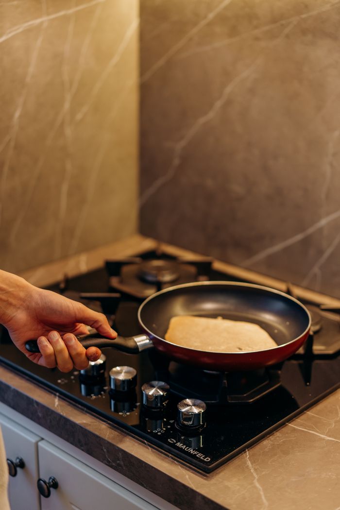 A close-up of a hand holding a frying pan with a pancake on a gas stove in a modern kitchen setting.